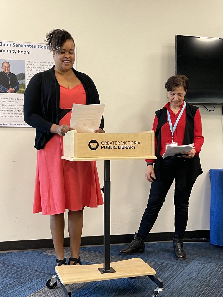Rachael Otukol (BCBHAS) and Naomi Charaf (GVPL) standing at the podium. Rachel is wearing a pink dress and Naomi is wearing black pants and a red shirt.